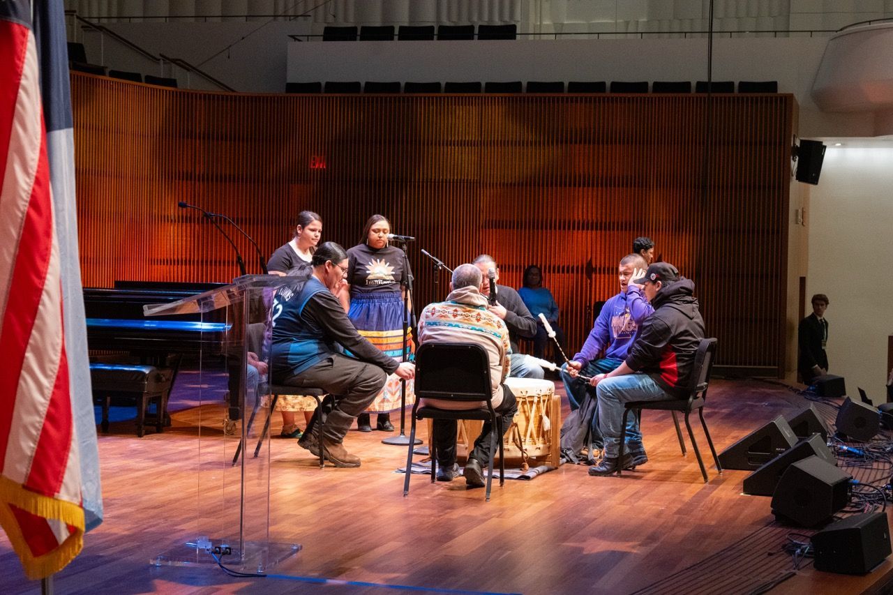 A group of eight people, including two women standing and six people seated in a circle on chairs, perform a traditional drum circle in a wooden-floored auditorium with an American flag and a lectern nearby.